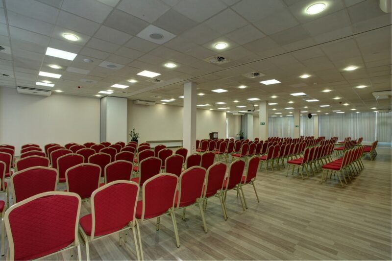 Duża sala konferencyjna, zastawiona kilkudziesięcioma krzesłami, ustawionymi w równe rzędy.
