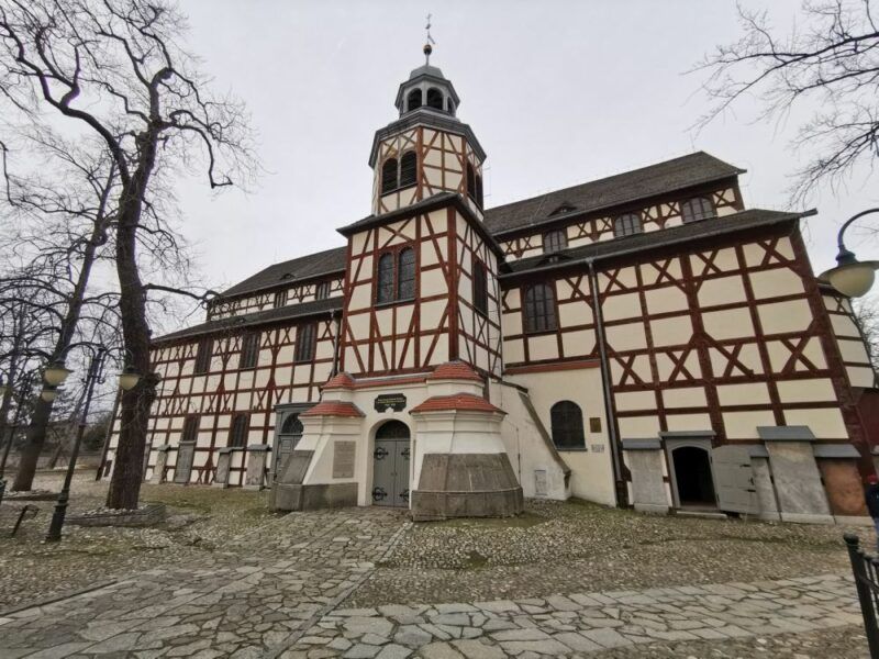 Na zdjęciu widoczny jest Kościół Pokoju w Jaworze, charakterystyczna budowla wpisana na listę UNESCO. Fasada kościoła wyróżnia się biało-brązowym układem drewnianych belek oraz centralną wieżą z barokowym hełmem. Otaczają go stare, bezlistne drzewa oraz brukowana ścieżka prowadząca do wejścia.