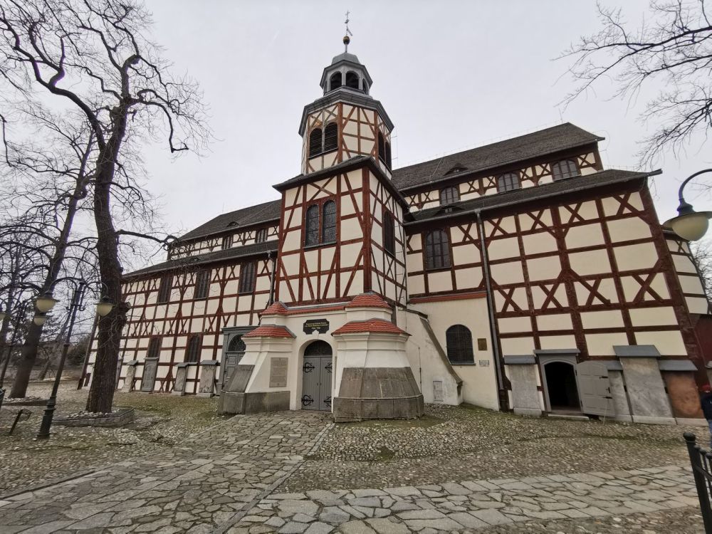 Na zdjęciu widoczny jest Kościół Pokoju w Jaworze, charakterystyczna budowla wpisana na listę UNESCO. Fasada kościoła wyróżnia się biało-brązowym układem drewnianych belek oraz centralną wieżą z barokowym hełmem. Otaczają go stare, bezlistne drzewa oraz brukowana ścieżka prowadząca do wejścia.