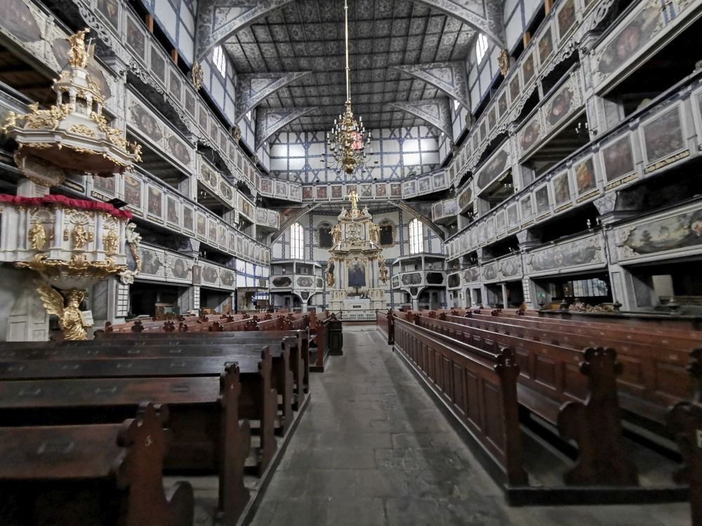 Zdjęcie przedstawia wnętrze Kościoła Pokoju w Jaworze, które jest duże i bogato zdobione. Wysokie balkony otaczają główną część kościoła, a na końcu widać piękny ołtarz. Po lewej stronie znajduje się złocona ambona, a z sufitu zwisa duży żyrandol.