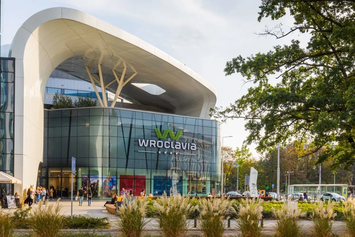 zdjęcie galerii handlowej Wroclavia o zachodzie słońca, sama galeria znajduje się na drugim planie a przed znajduję się pięć rownolegle ułożonych krzaków oraz po lewej jest wysokie drzewo