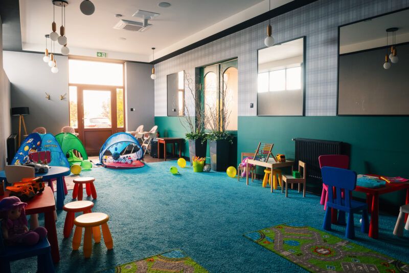 Duża przestronna sala zabaw, z dużą ilością stolików i krzesełek dziecięcych, małych namiotów oraz innych zabawek.