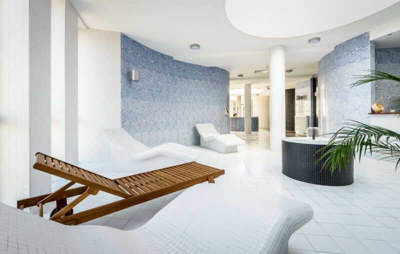 spa z drewnianymi oraz białymi, zaokrąglonymi leżakami. całośc jest wykonania estetycznie w białych kolorach. na dalekim planie znajduję się jacuzzi