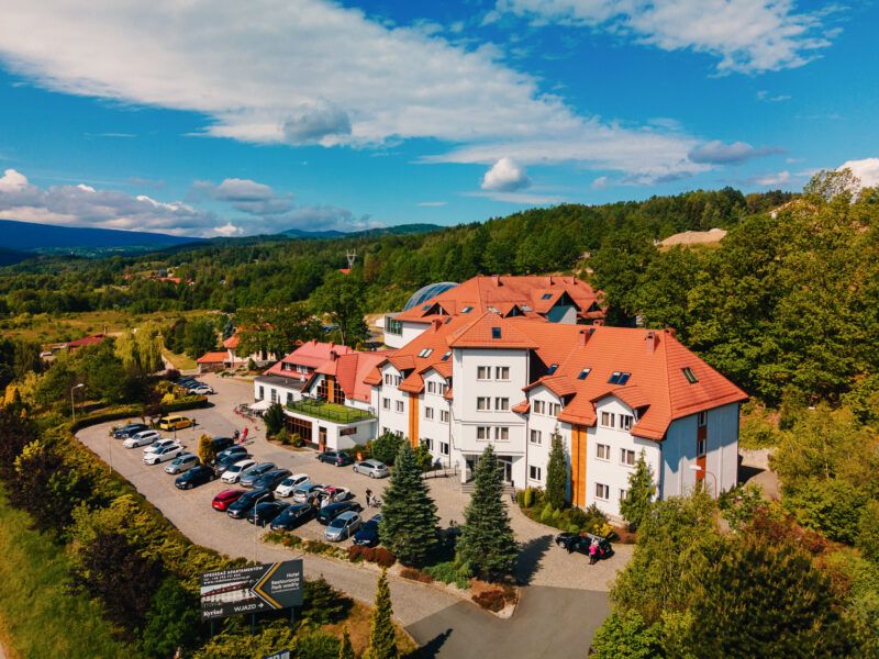 Zdjęcie z drona przedstawiające budynek hotelowy na tle górskiego krajobrazu. Długi, trójkondygnacyjny budynek z wejściem wyszczególnionym symetrycznym, trójkątnym wykuszem. Przed budynkiem znajduje się duży parking.