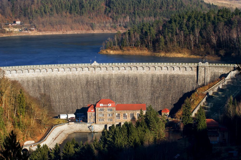Widok z lotu ptaka, na kamienną zaporę na rzece. W tle las.
