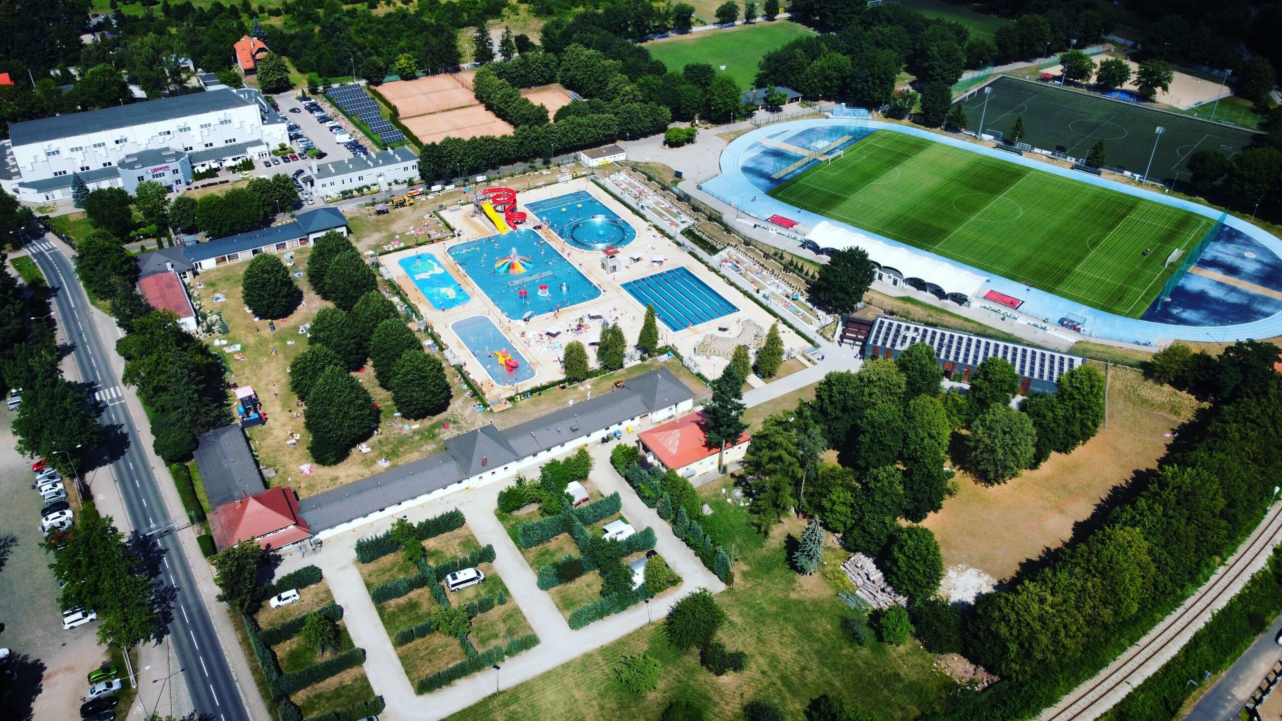 Widok z drona na kompleks sportowo-rekreacyjny przedstawiający między innymi pole campingowe, boisko sportowe oraz baseny zewnętrzne z brodzikiem dla dzieci, podwójną zjeżdżalnią i wirem wodnym. 