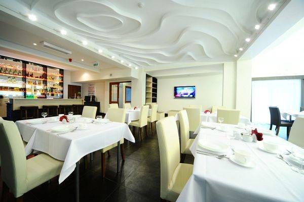 Duża, jasna sala restauracyjna z prostokątnymi stołami, nakrytymi białymi obrusami i fotelami z kremowym obiciem.