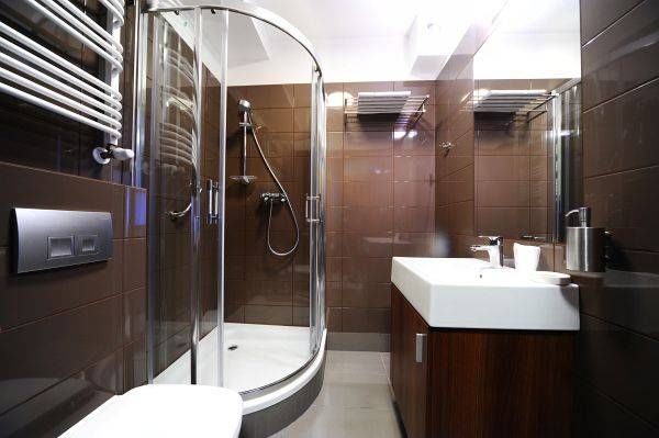 Łazienka hotelowa. Narożna kabina prysznicowa, prostokątny zlew na szafce, duże lustro oraz podwieszany sedes. Na ścianach ciemne kafelki.