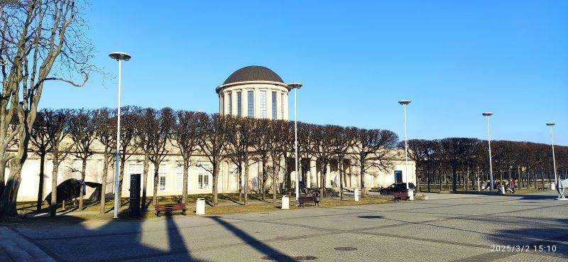 Modernistyczny budynek o prostych, geometrycznych kształtach, wyróżniający się czterema charakterystycznymi kopułami na dachu, otoczony przestronnym terenem i subtelną fasadą z czerwonej cegły.