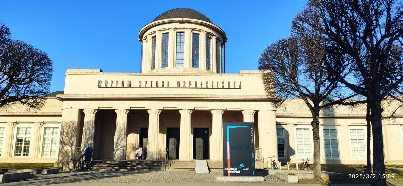 Modernistyczny budynek o prostych, geometrycznych kształtach, wyróżniający się czterema charakterystycznymi kopułami na dachu, otoczony przestronnym terenem.