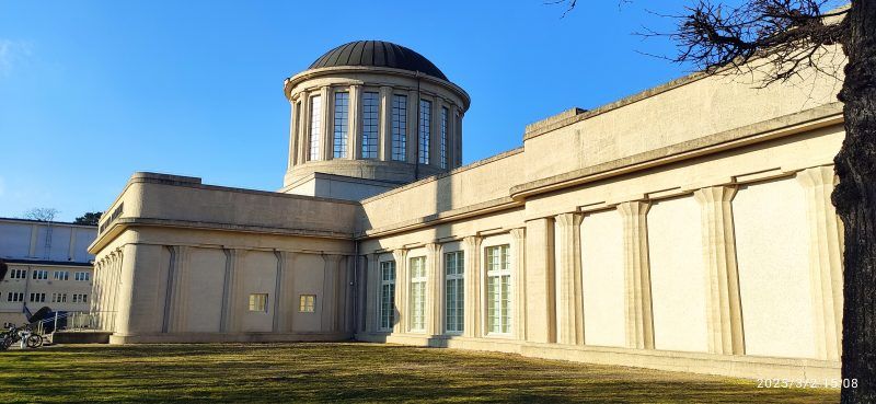 Modernistyczny budynek o prostych, geometrycznych kształtach, wyróżniający się czterema charakterystycznymi kopułami na dachu, otoczony przestronnym terenem.