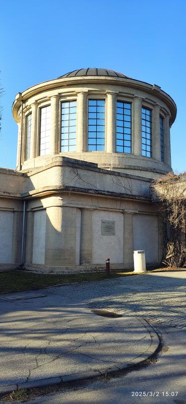 Modernistyczny budynek o prostych, geometrycznych kształtach, wyróżniający się czterema charakterystycznymi kopułami na dachu, otoczony przestronnym terenem.