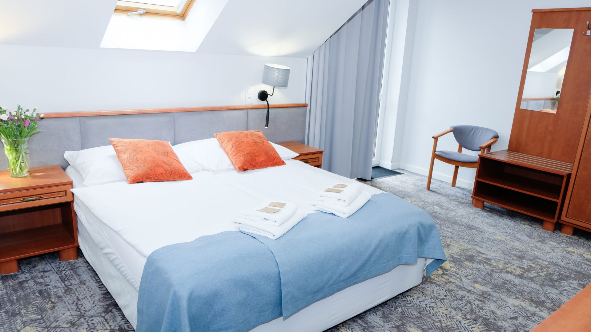 Pokój hotelowy z dużym podwójnym łóżkiem, szafkami nocnymi, szafką z lustrem. Na podłodze przytulną wykładziną.