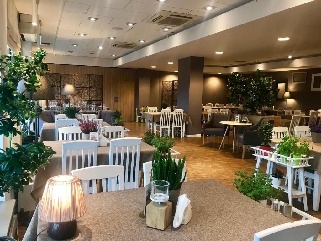 Przestronne, eleganckie wnętrze restauracji z białymi drewnianymi krzesłami oraz prostokątnymi stołami nakrytymi obrusami. Dekorację stanowią małe lampki, dużo 