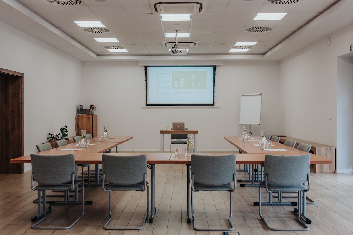 Sala konferencyjna z podłużnymi stołami ułożonymi w literę U i przylegającym doń krzesłami. Na ścianie ekran multimedialny.