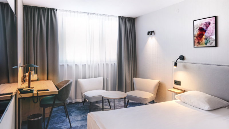 Elegancki pokój hotelowy. Okno przesłonięte długimi zasłonami szafka z lampką i fotelem, mały komplet wypoczynkowy oraz łóżko