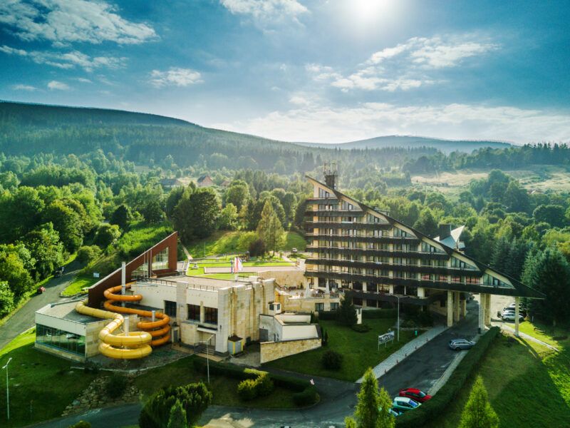 Duży budynek hotelowy na tle lasu oraz gór. Budowla w kształcie trójkąta z przylegającym doń kompleksem sportowo-rekreacyjnym z basenem. Uwagę przyciągają duże zjeżdżalnie.