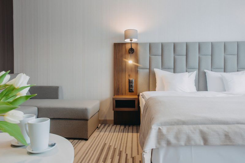 Nowoczesny, pokój hotelowy z podwójnym łóżkiem z zagłówkiem obitym tkaniną. Na podłodze wykładzina. Widoczny kawałek kolistego stolika i sofy.