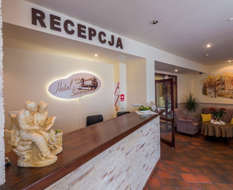 Recepcja hotelowa. Wysoka lada zakończona drewnem. Na ścianie logo i nazwa hotelu.