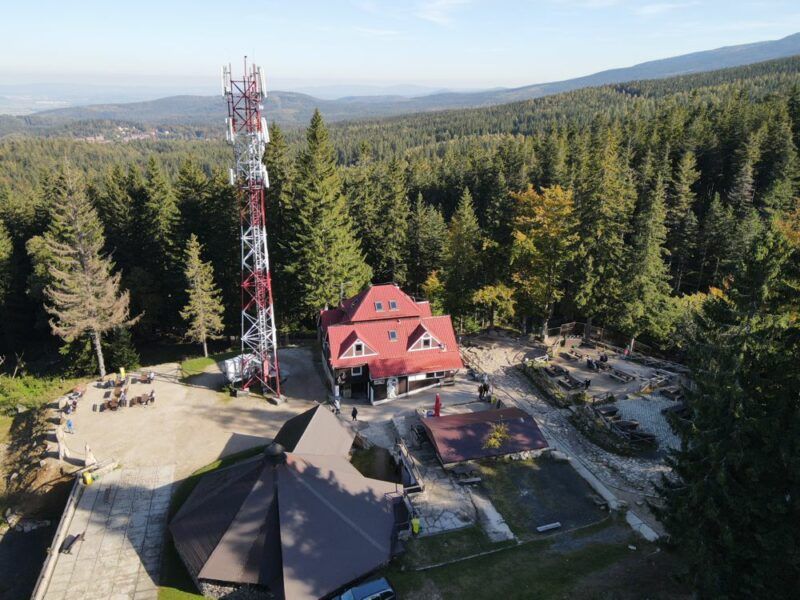 Zdjęcie z drona przedstawiające rozległy plac w lesie iglastym, na terenie którego ustawiony jest budynek schroniask, duża wiata oraz maszt nadajnikowy. 