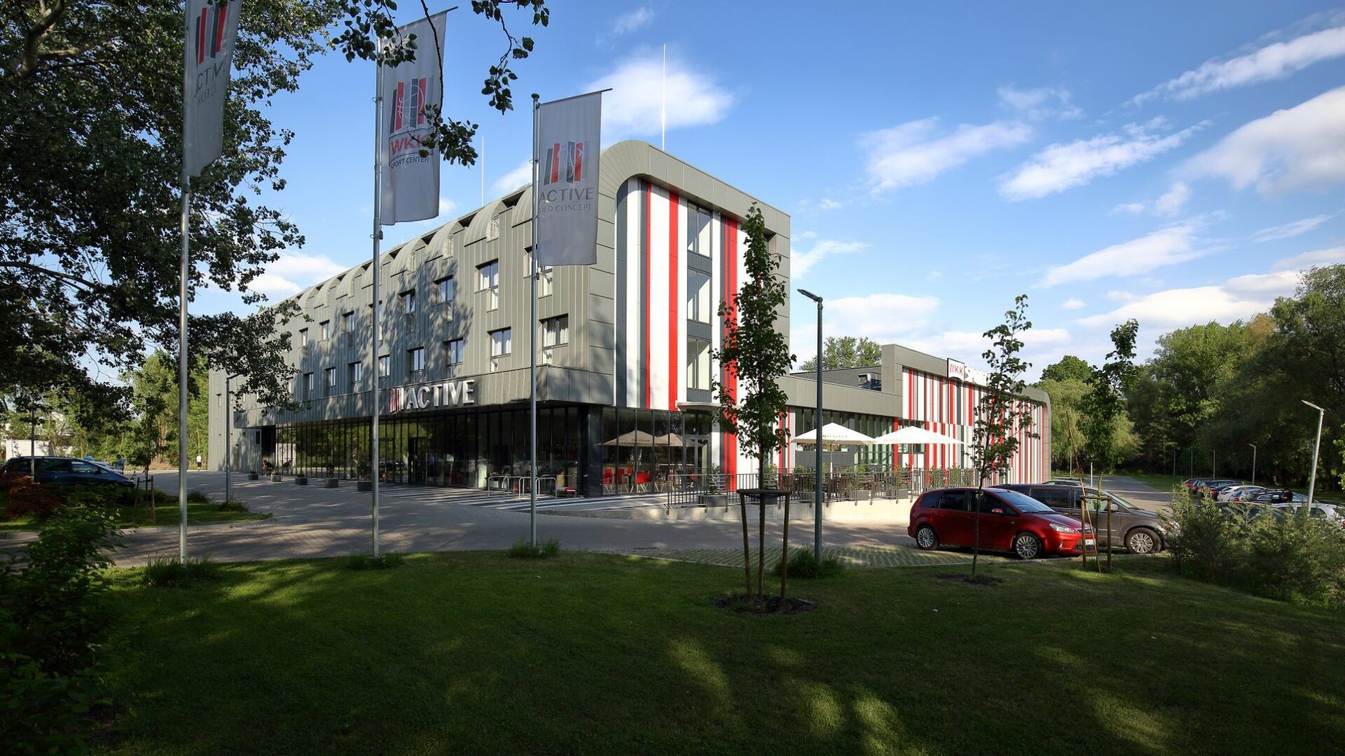 Nowoczesny, czterokondygnacyjny, podłużny budynek, zdobiony kolorowymi pionowymi pasami. Przed budynkiem parking oraz stelaże z flagami z logo i nazwą hotelu.