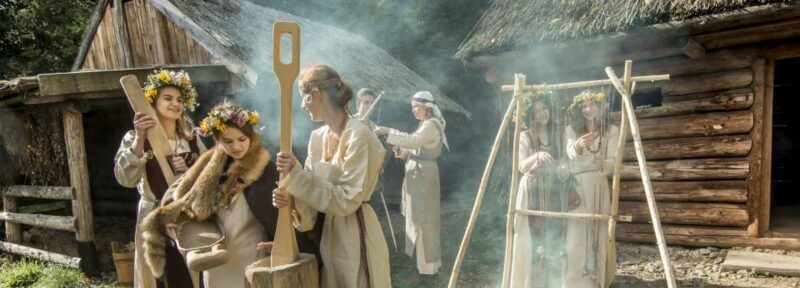 Zdjęcie przedstawia grupę kobiet w słowiańskich strojach w otoczeniu wiejskich, drewnianych chatek.