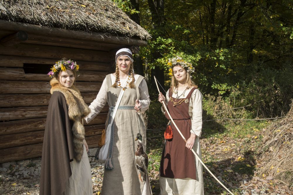 Zdjęcie przedstawia grupę kobiet w słowiańskich strojach w otoczeniu wiejskich, drewnianych chatek.