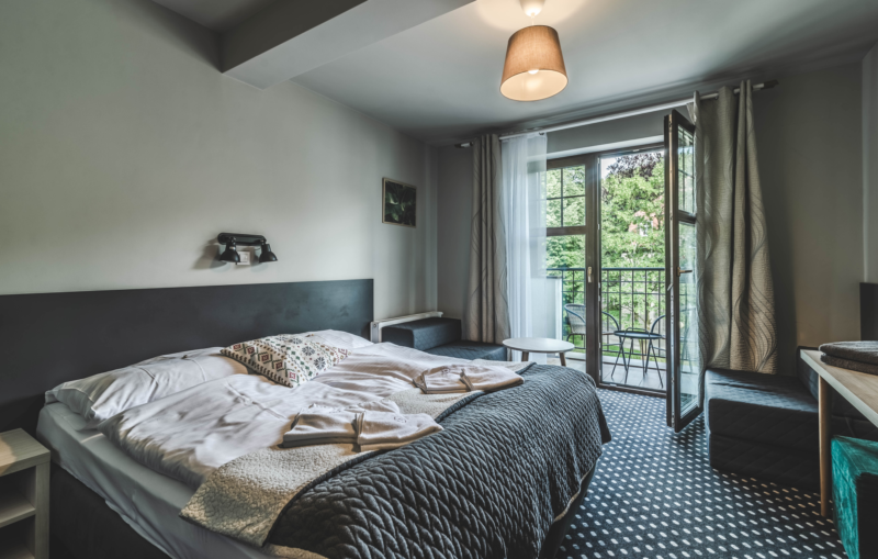 Pokój hotel z podwójnym łóżkiem. Szerokie okno prowadzi na balkon.