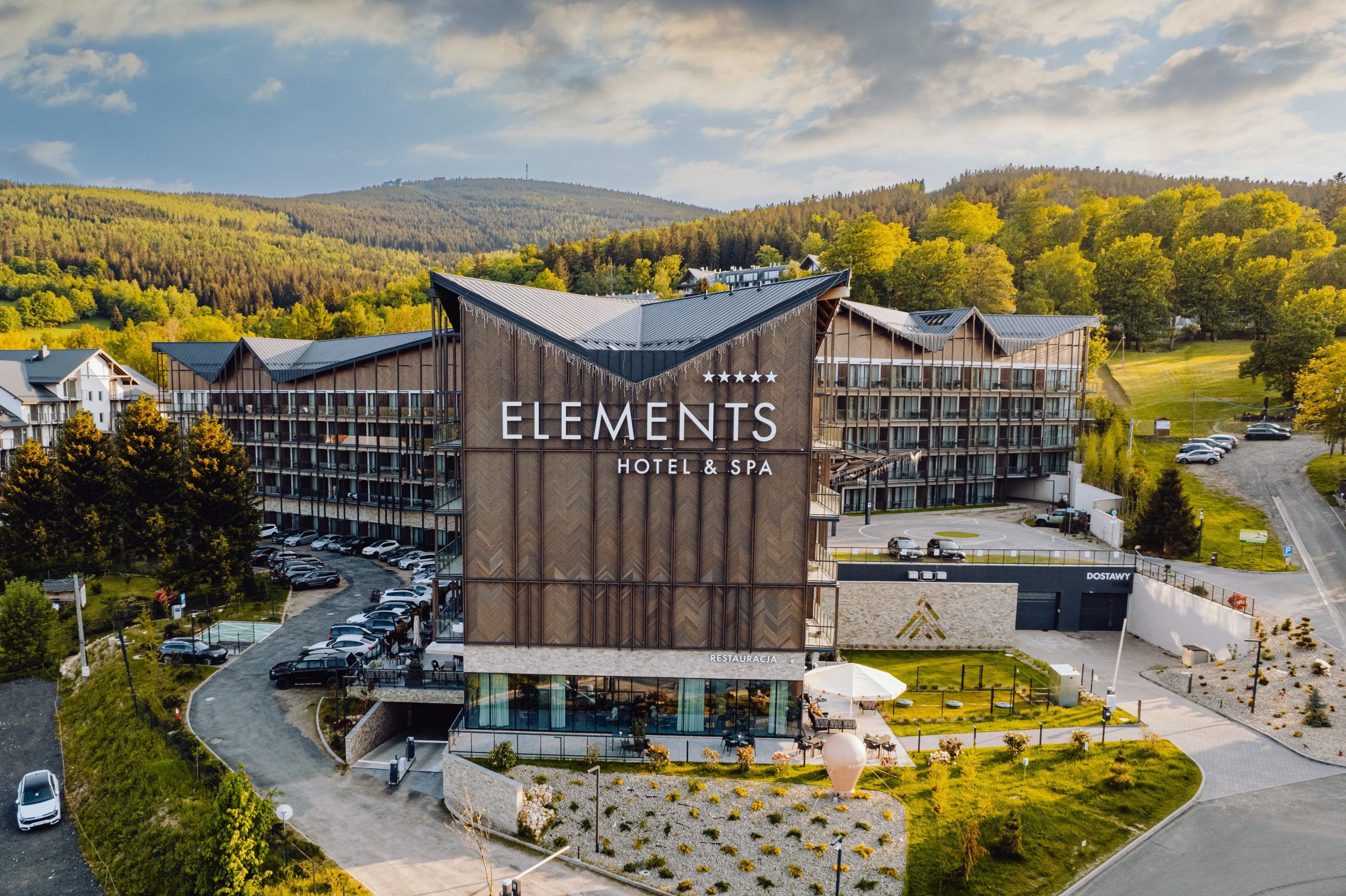 Zdjęcie z drona z dużym budynkiem hotelowym, na szczycie elewacji z elementami drewnianymi logo i nazwa hotelu Elements. Przed budynkiem elementy infrastruktury zielonej, parking. za budynkiem widok na las i góry.