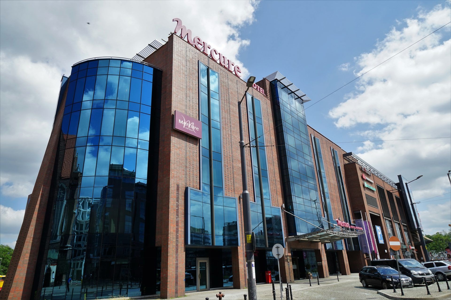 Nowoczesny budynek z fasadą ze szkła oraz materiałów imitujących cegłę. Bryła nieregularna. Na szczycie oraz elewacji logo i nazwa hotelu. 