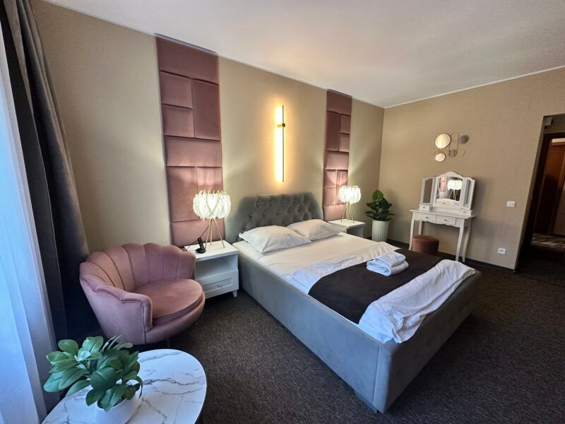 Elegancki pokój hotelowy, z dużym podwójnym łóżkiem, elegancką toaletką, lampkami nocnymi oraz fotelem.