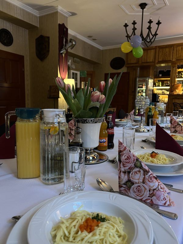 Restauracja hotelowa. Zbliżenie na stół nakryty obrusem. Na środku wazon z kwiatami oraz dzbanki z napojami oraz talerze z makaronem na rosół.