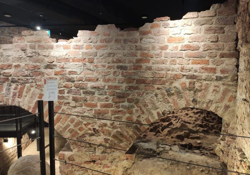 Fragment starego muru z cegły z łukiem, prawdopodobnie część fortyfikacji. Jest dobrze oświetlony i zabezpieczony barierkami.