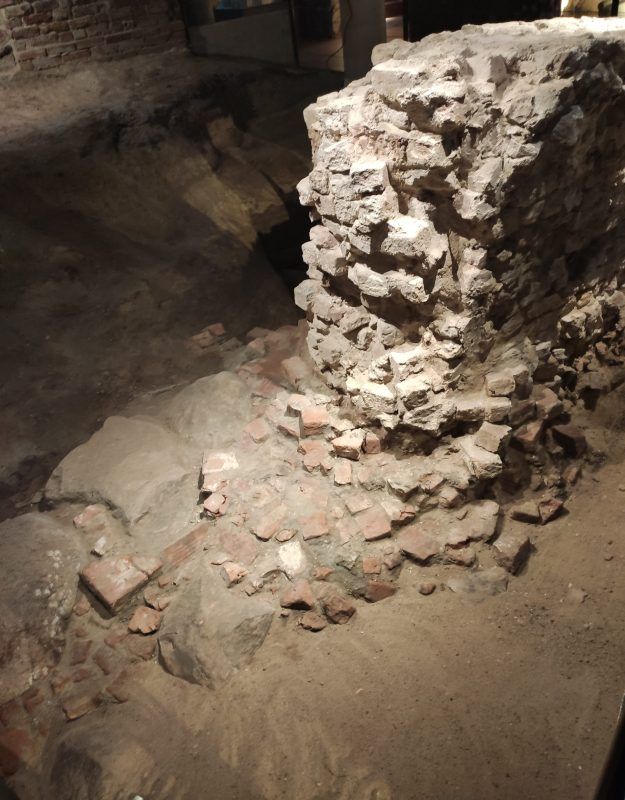 Ruiny z cegieł i kamieni, częściowo zasypane piaskiem. To pozostałości dawnej konstrukcji odsłonięte podczas badań archeologicznych.