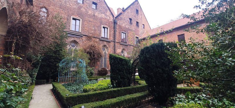 spokojny, zadbany ogród otoczony ceglaną zabudową, z przyciętymi żywopłotami i dekoracyjną żelazną arką.