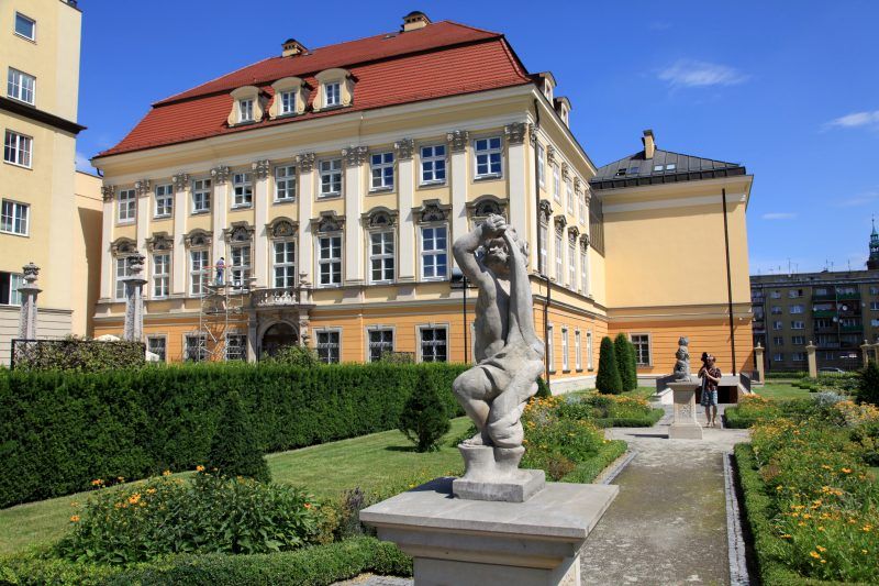 Na pierwszym planie widać ścieżkę, wzdłuż której stoją pomniki a w oddali widać zabytkowy budynek w kolorze pastelowo żółtym, w którym znajduje się muzeum.