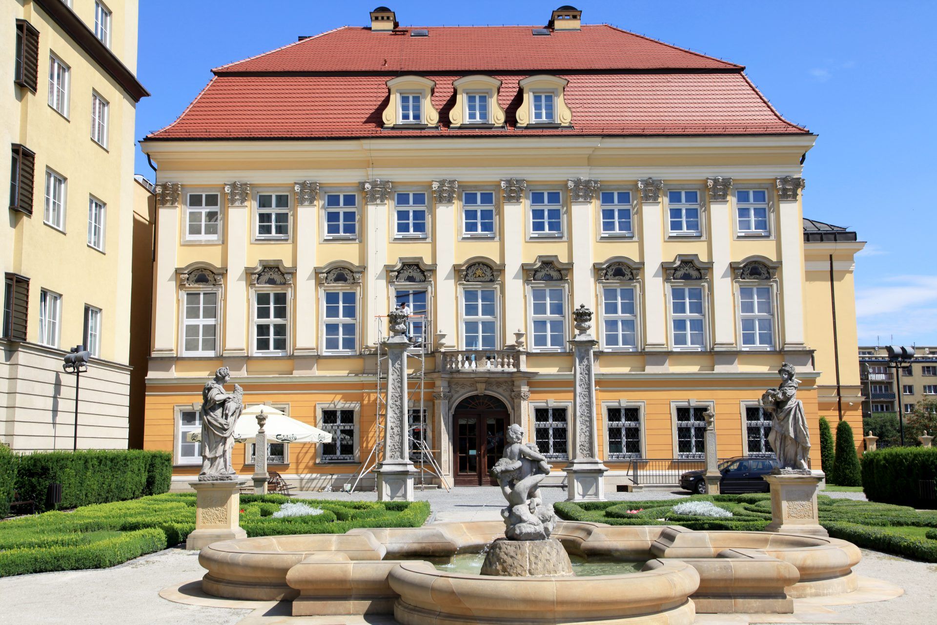 Zabytkowy budynek, w którym znajduje się wystawa muzealna w kolorach żółtych z pięknymi oknami na tle jasnego nieba. Nieopodal wejścia widać zabytkową fontannę