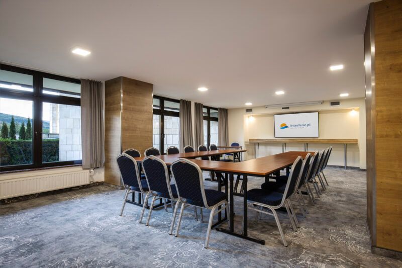 Sala konferencyjna z długim drewnianym stołem ułożonym w literę U oraz przylegającymi doń krzesłami biurowymi. Na ścianie wisi ekran multimedialny. 