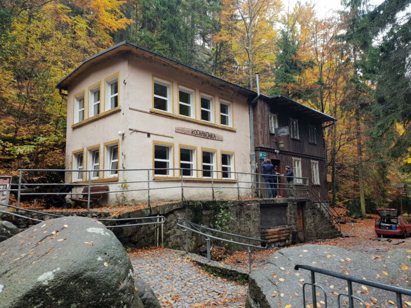 Niewielki prostokątny, dwupiętrowy budynek schroniska na tle jesiennego lasu.