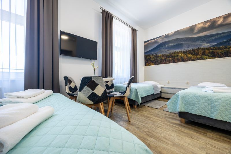 Pokój hotelowy z 3 pojedynczymi łóżkami. Na ścianie fototapeta z górskim krajobrazem. Na prostopadłej ścianie telewizor oraz stolik z krzesłami.