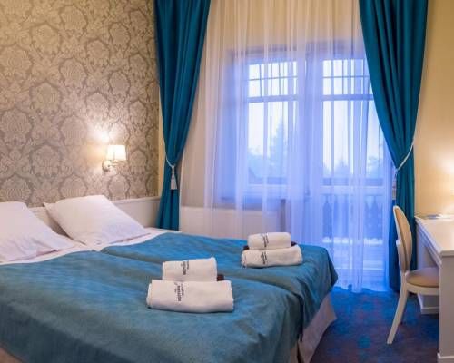 Pokój noclegowy w beżowo-błękitnej tonacji. Długie zwisające, podwiązane zasłony, łóżko podwójne oraz komoda z krzesłem.