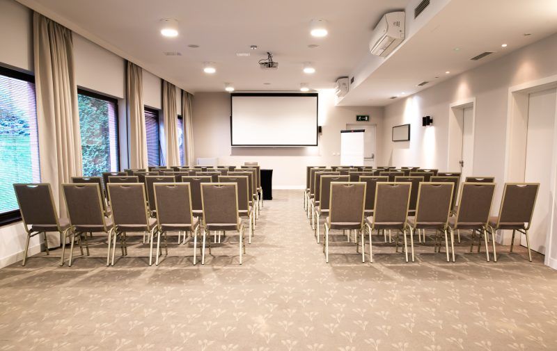 Sala konferencyjna z kilkunastoma rzędami krzeseł. Na przodzie duży ekran multimedialny.