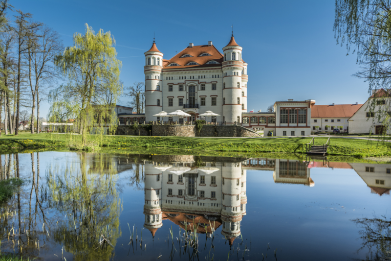Widok na zabytkowy pałac z wieżyczkami w narożach z widocznym stawem na pierwszym planie
