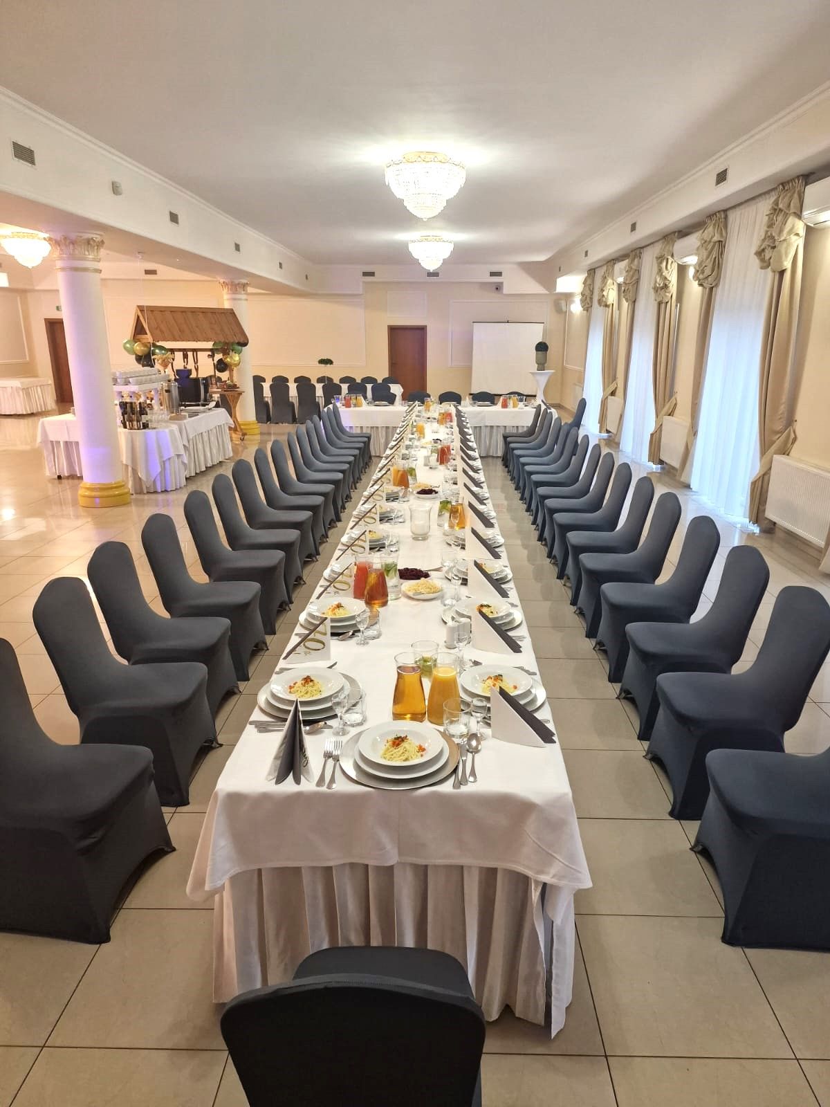 Duża sala restauracyjna (balowa), przyszykowana na uroczystość. Długi stół z kilkudziesięcioma krzesłami po obu stronach. Stół nakryty, w talerzach makaron na rosół.