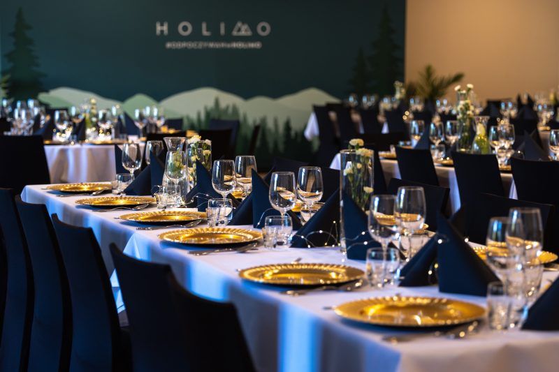 Sala restauracyjna. Zbliżenie na podłużny stół, nakryty obrusem i zastawiony eleganckimi talerzami, kieliszkami oraz serwetkami. Dekorację stanowią drobne lampki oraz goździki w wazonach. Na ścianie logo i nazwa hotelu Holimo.
