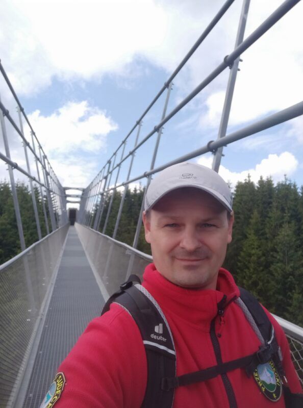 mężczyzna w czerwonym polarze z odznaką przewodnika sudet, idący przez szary most