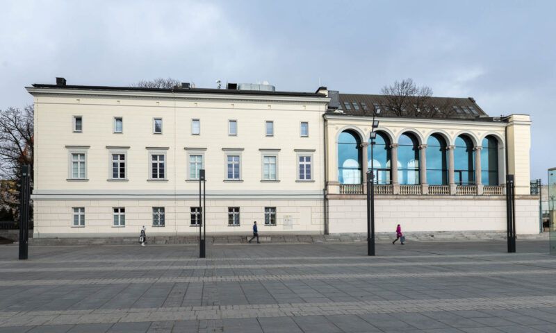 Zdjęcie przedstawia budynek Muzeum Teatru. Przed nim znajduje się deptak, a sam budynek jest w kształcie prostopadłościanu, z tego lewa jego część posiada liczne, ustawione symetrycznie obok siebie okna, a druga prawa część, składa się z sześciu dużych okien, które u górnej swojej części są zaokrąglone.