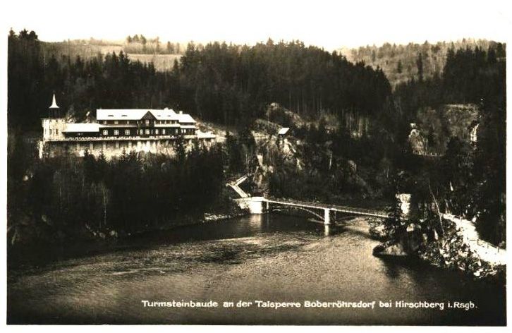 stare, czarno-białe zdjęcie w słabej jakości, ukazujące panoramę zawierającą w sobie duże schronisko oraz most znajdujący się z jego prawej strony