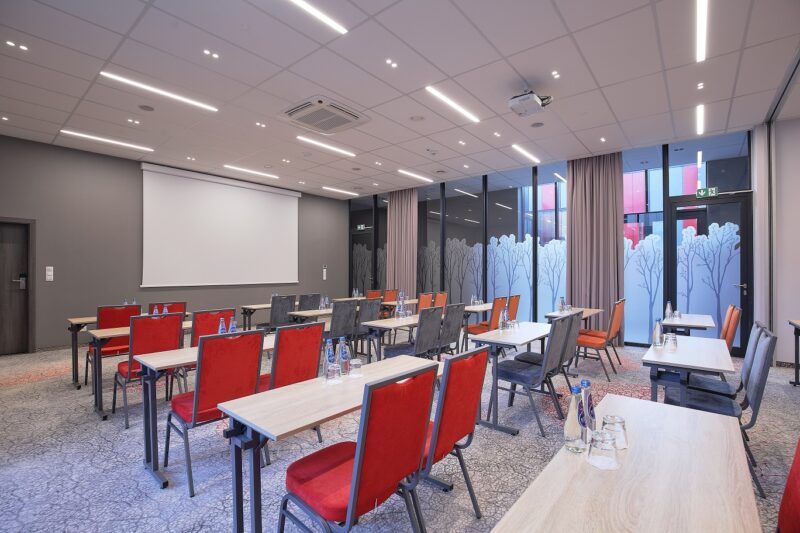 Sala konferencyjna z 4 rzędami prostokątnych stołów. Na ścianie duży ekran.