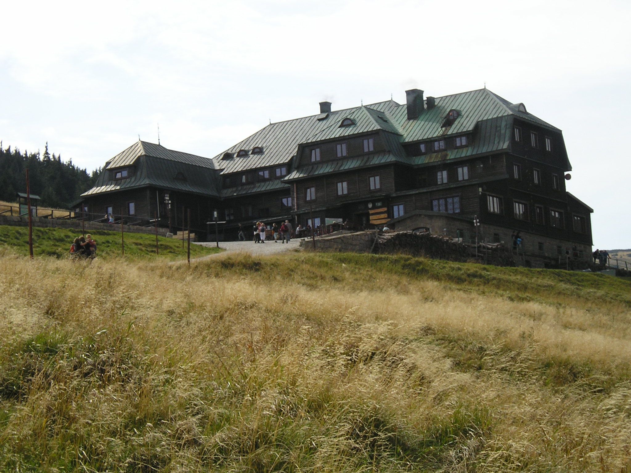 widok od dołu na schronisko w pogodny dzień, przed obiektem znajduję się łąka, a sam budynek jest zrobiony z drewnianej konstrukcji, a w niej wbudowane są liczne małe okna